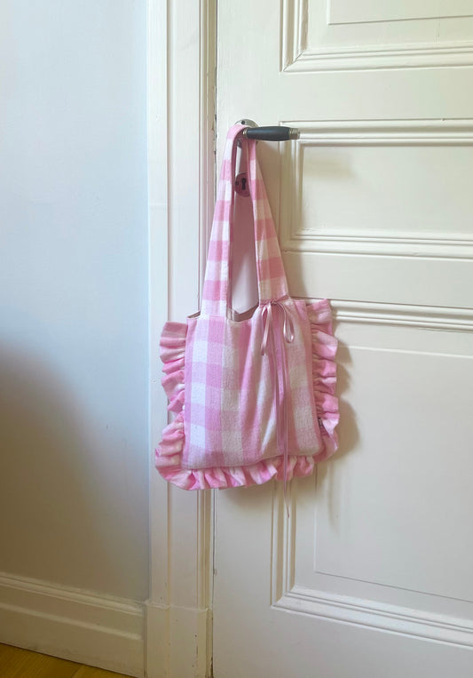 White and pink checkered tote bag with decorative ruffles. Hanging on a door handle.